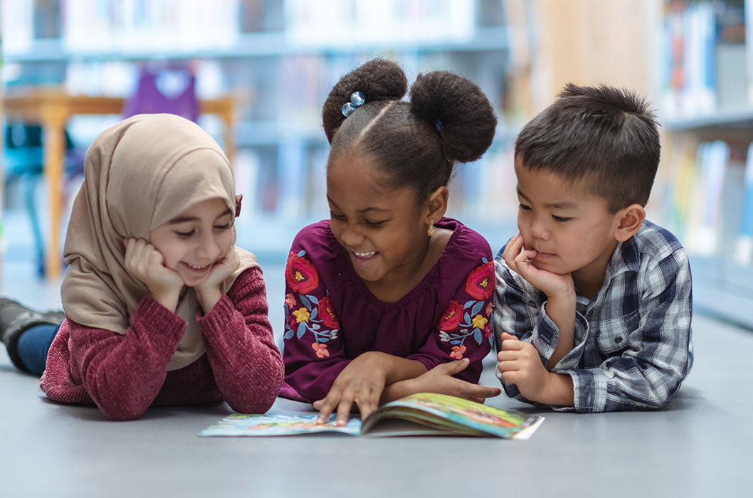 Three (3) kids who are friends are laying on the floor in between bookshelves in the library. They are reading a book together. They are all smiling and enjoying their afternoon.